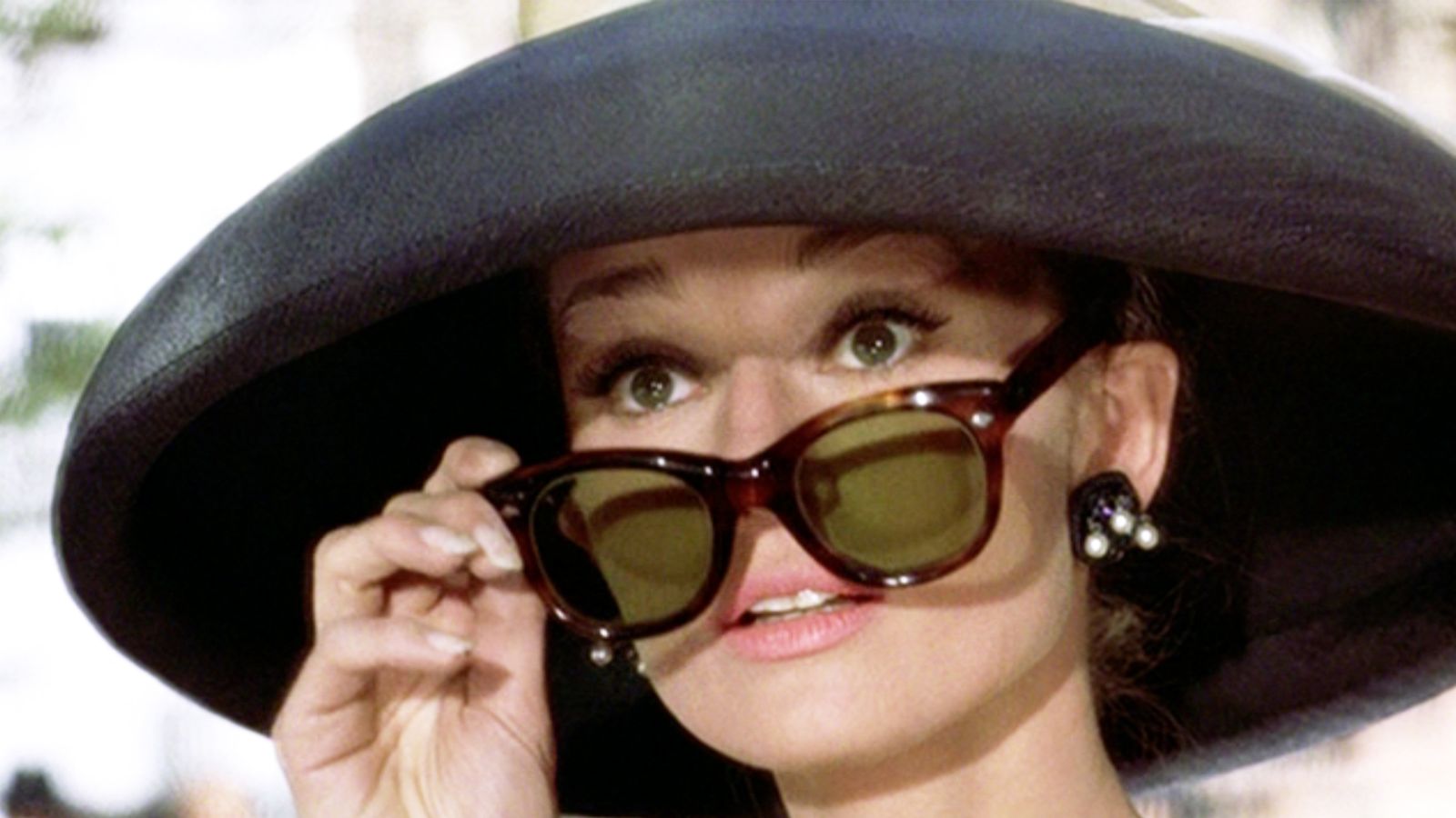 Audrey Hepburn jako Holly Golightly ve filmu Snídaně u Tiffanyho, v širokém klobouku a želvovinových slunečních brýlích Manhattan